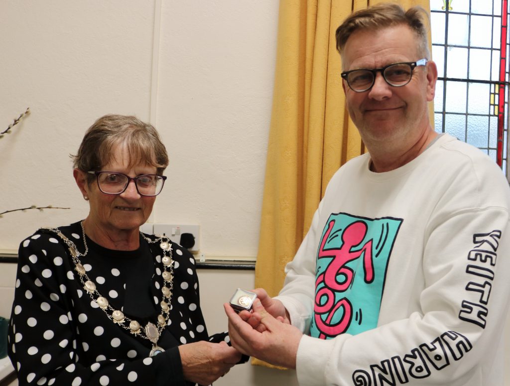 A woman in chains of office hands an enamel badge to a man.