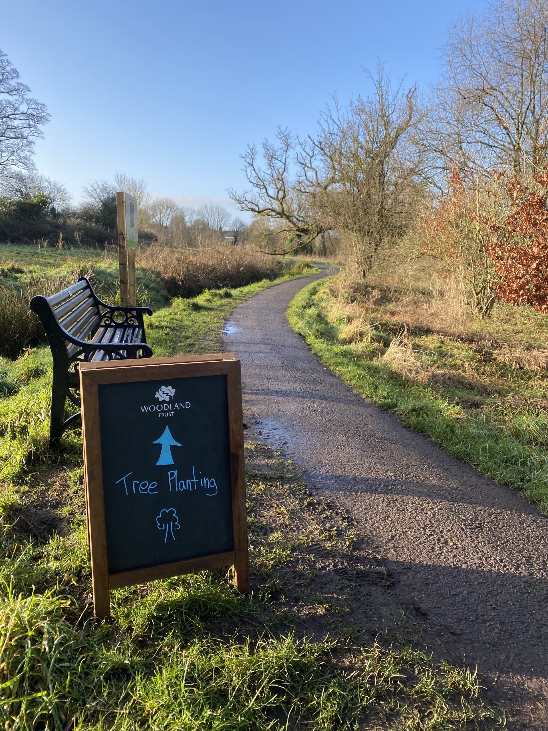A chalkboard sign pointing into a field. The board has The Woodland Trust logo and says "Tree Planting"
