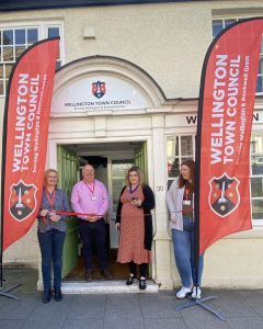 A group of people cut a ribbon outside of a building with Wellington Town Council logos and signage.