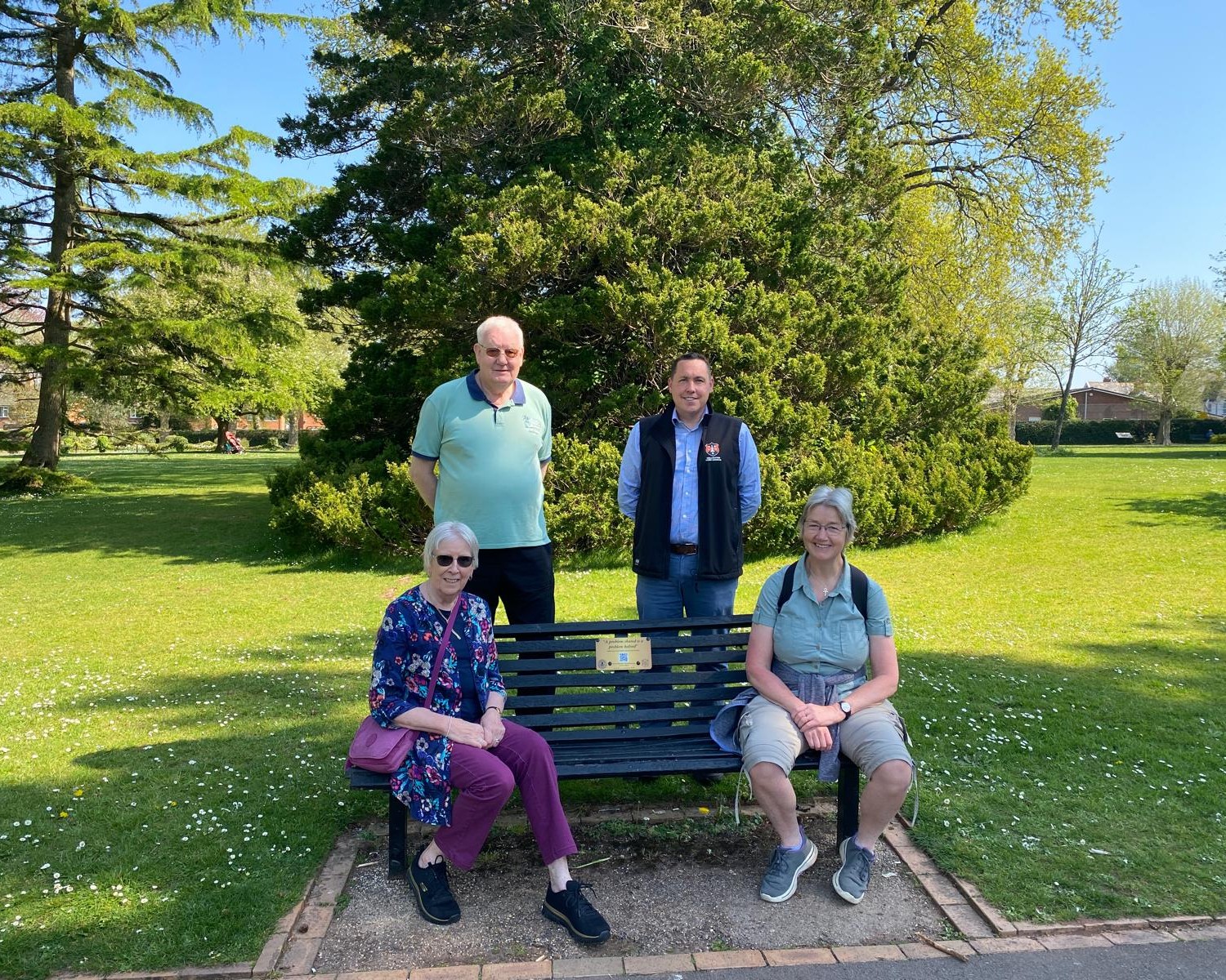 A group of people surround a bench with a plaque that reads "A Problem Shared is a Problem Halved".